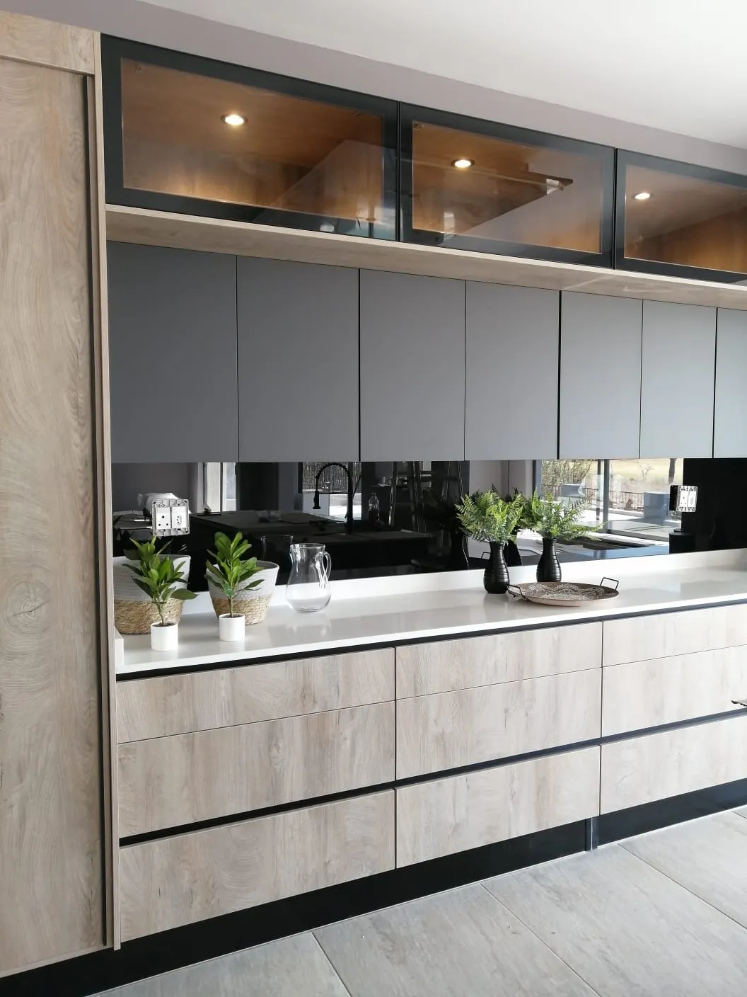 Light oak and matte grey kitchen with lit glass display cabinets above a black mirror splashback, by N.D.I.